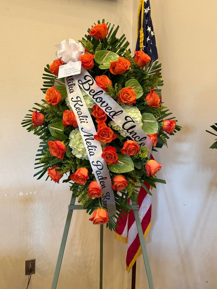 A standing spray on an easel of orange roses, white hydrangea and green anthurium with a hand lettered sash.