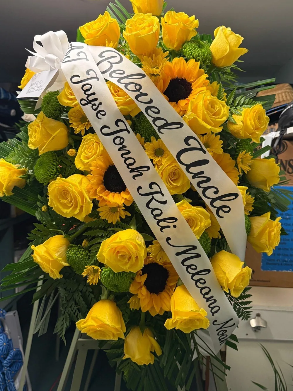 Close view of yellow roses and greenery beside a white ribbon sash lettered by hand.