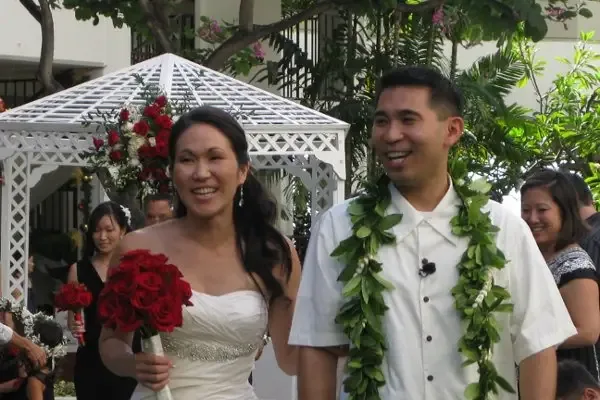 A bride and groom standing at a white gazebo, the groom wearing a maile lei and the bride holding a red rose bouquet.