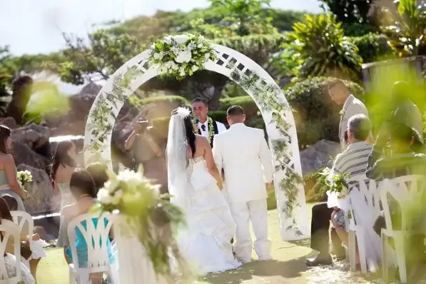 A couple at a white ceremony arch dressed with greenery and white flowers.